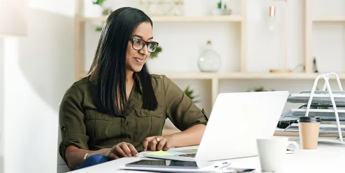 Woman with glasses in olive green shirt working on laptop at desk in bright modern office space with coffee cup nearby.