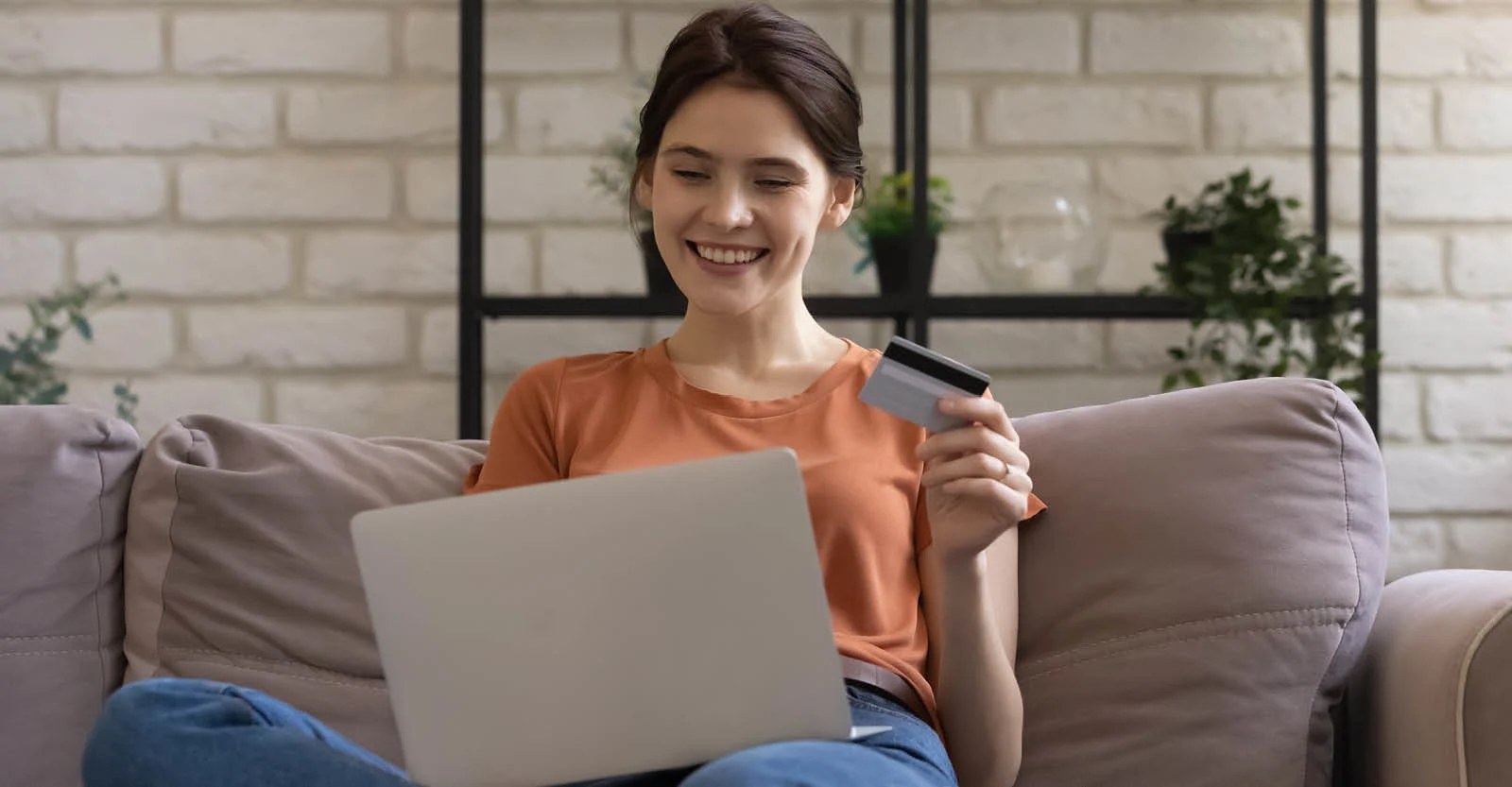 Woman in orange shirt sitting on gray couch, smiling while holding credit card and using laptop for online shopping.