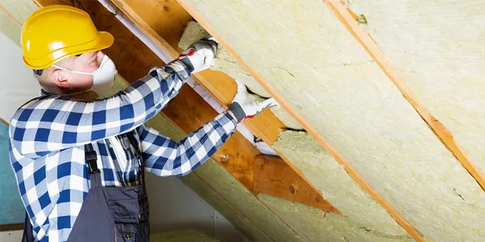 Worker in yellow hard hat and plaid shirt installing insulation between wooden roof beams from below.