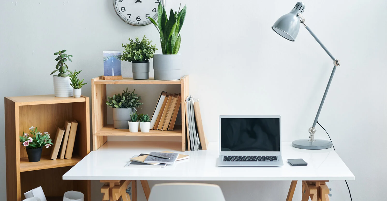 Modern minimalist home office with white desk, laptop, gray desk lamp, wooden shelving units with plants, and wall clock.