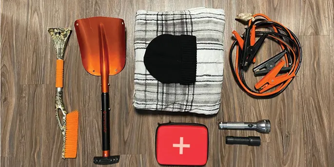 Winter car emergency kit with shovel, blanket, hat, first aid kit, flashlight, and jumper cables on wood surface.