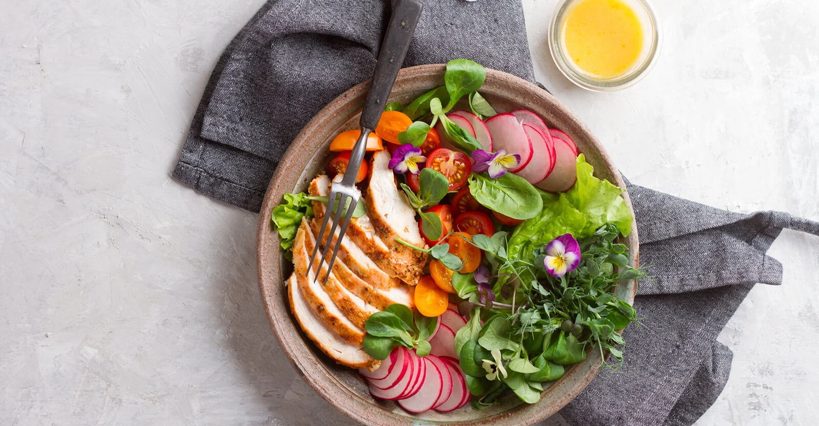 Colorful salad bowl with sliced grilled chicken, radishes, tomatoes, greens, and edible flowers on gray napkin.