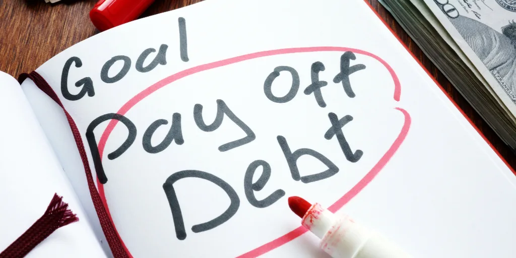 Notebook with handwritten text reading Goal Pay Off Debt circled in pink marker, with cash and red pen nearby.