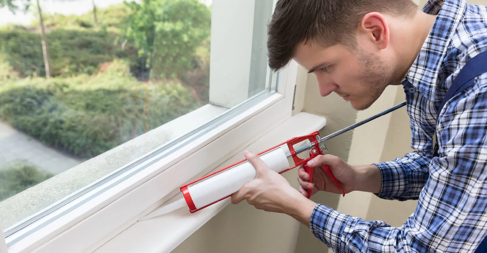 Man in blue plaid shirt applying white caulk with red caulking gun along window frame indoors with green foliage visible outside.