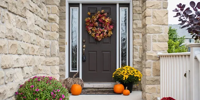 Brown front door with autumn wreath, flanked by glass panels, pumpkins and yellow mums on stone porch with fall decor.