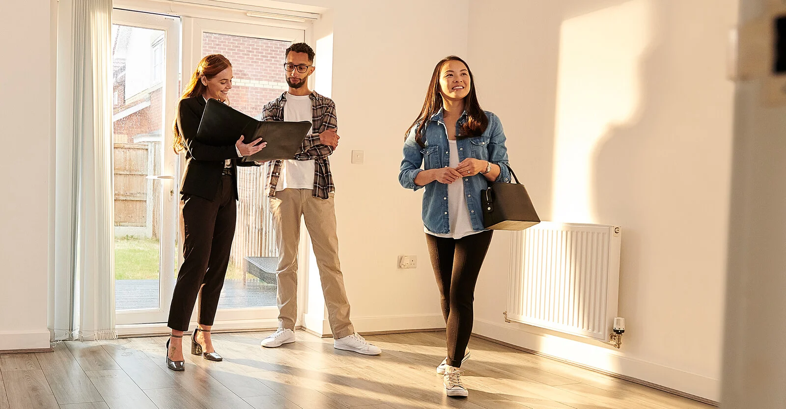 Real estate agent showing empty apartment to young couple viewing property with natural light from windows.