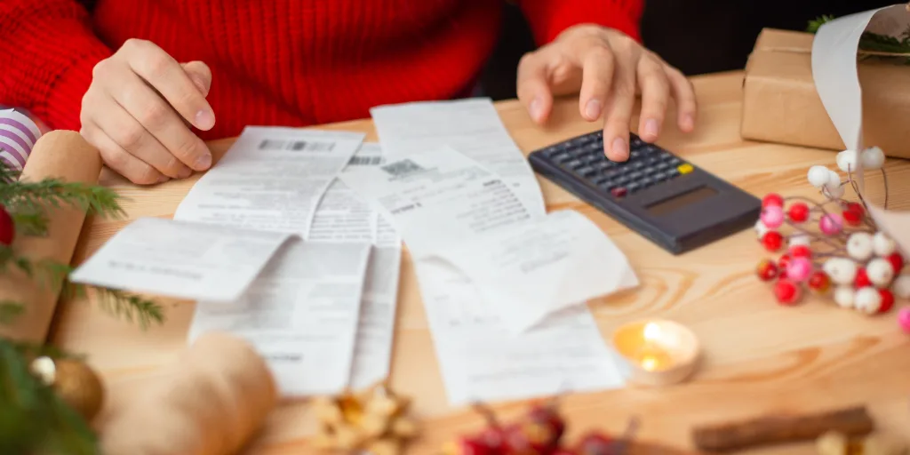 Person in red sweater using calculator while reviewing receipts on wooden table surrounded by holiday decorations and gifts.