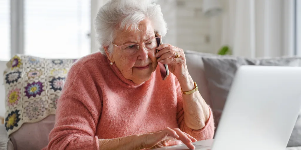 Senior woman with white hair and glasses wearing a coral sweater talking on phone while using laptop at home on couch.