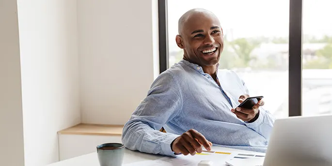 Smiling businessman in blue shirt holding phone at desk with laptop and coffee cup near bright window