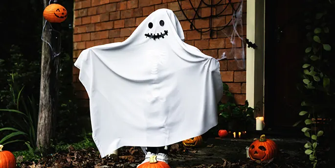 Child in white ghost costume with drawn smile stands on porch with jack-o'-lanterns and Halloween decorations.