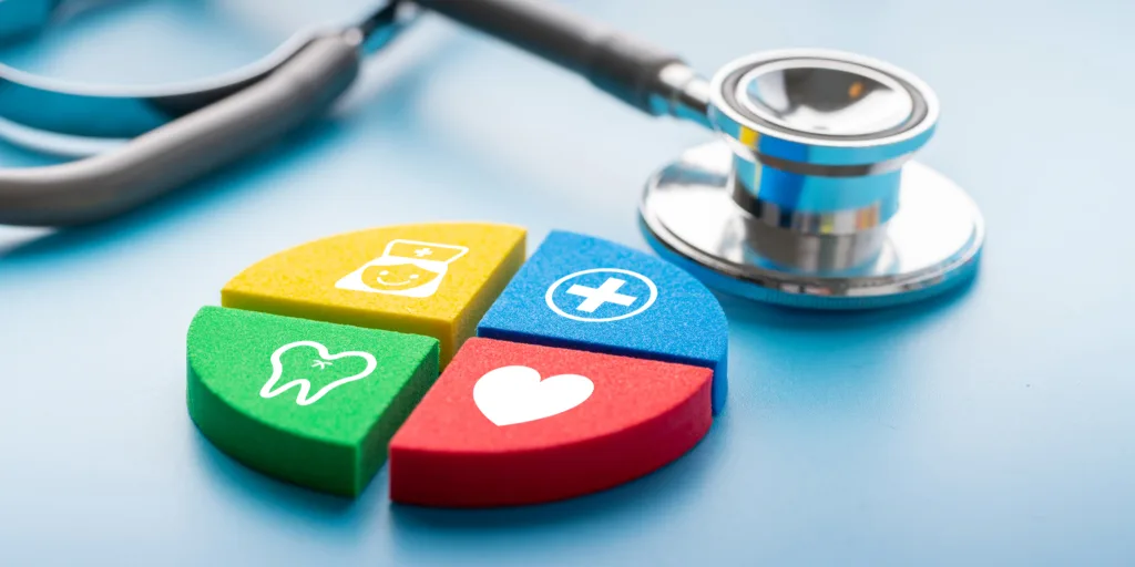 Stethoscope beside four colorful puzzle pieces with healthcare icons: tooth, medical bag, cross, and heart on blue surface.