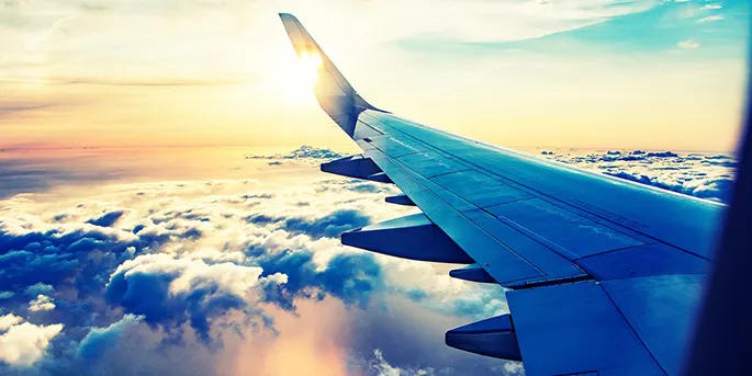 Airplane wing viewed from window seat flying above white clouds at sunset with golden sun rays illuminating the sky