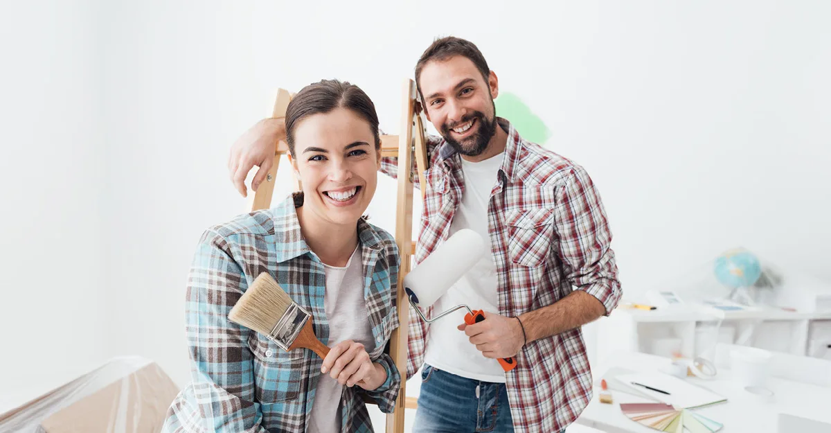 Smiling couple in plaid shirts holding paintbrush and paint roller during home renovation project