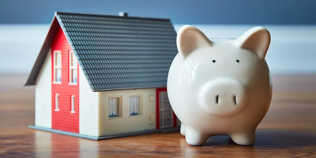 White piggy bank next to a miniature house model with red and white walls and gray roof on wooden surface.