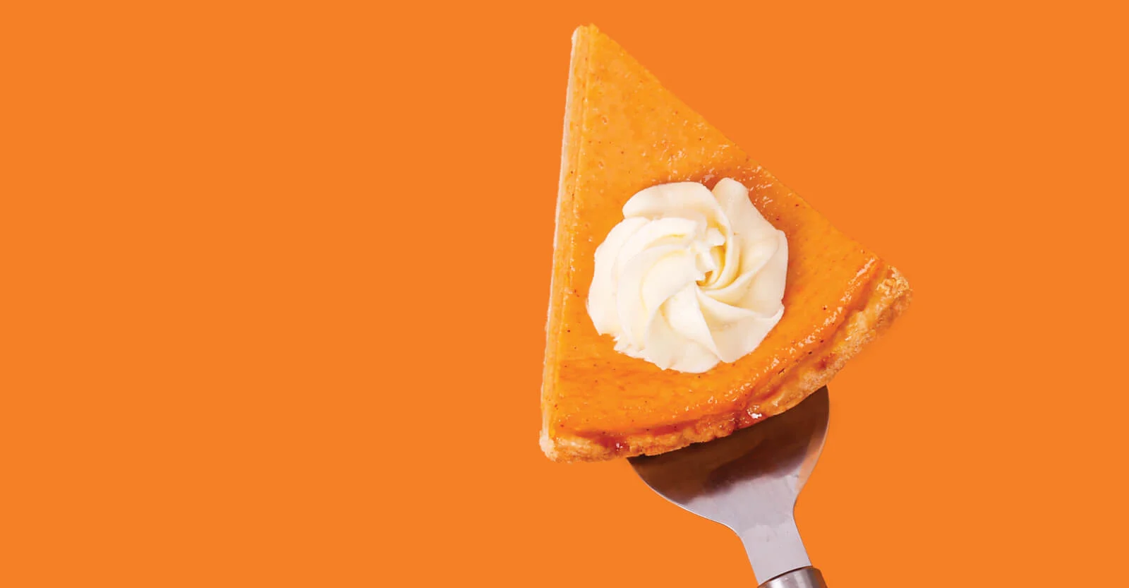 Slice of pumpkin pie topped with whipped cream on a silver spatula against an orange background.