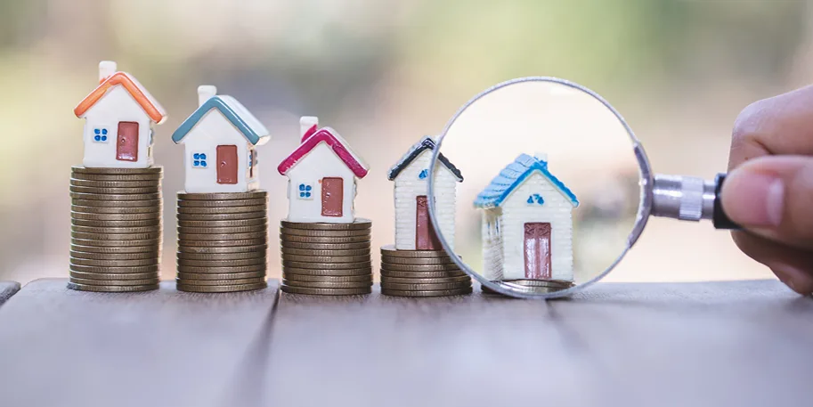 Miniature houses on stacks of coins with a magnifying glass examining one house, representing property investment analysis.