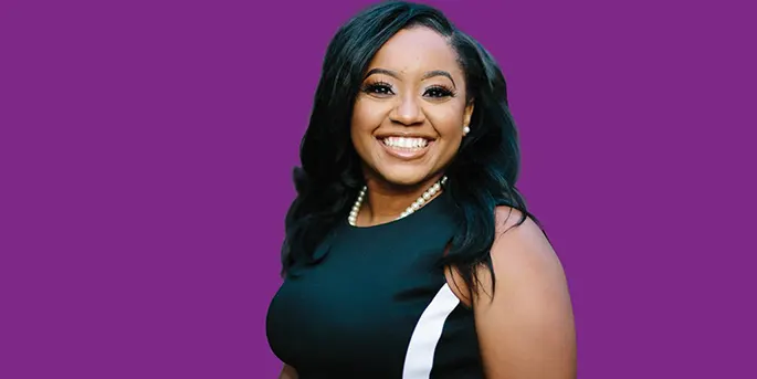 Smiling woman with long dark hair wearing a black dress and pearl necklace against a vibrant purple background.
