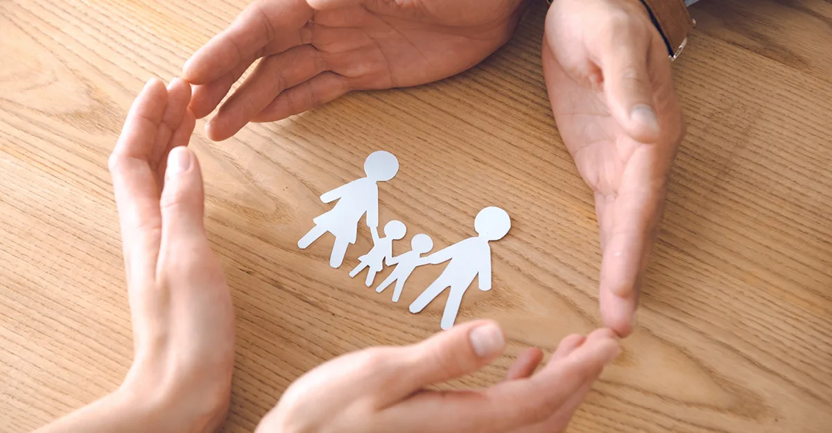 Hands forming protective circle around white paper cutout family figures on wooden table surface.