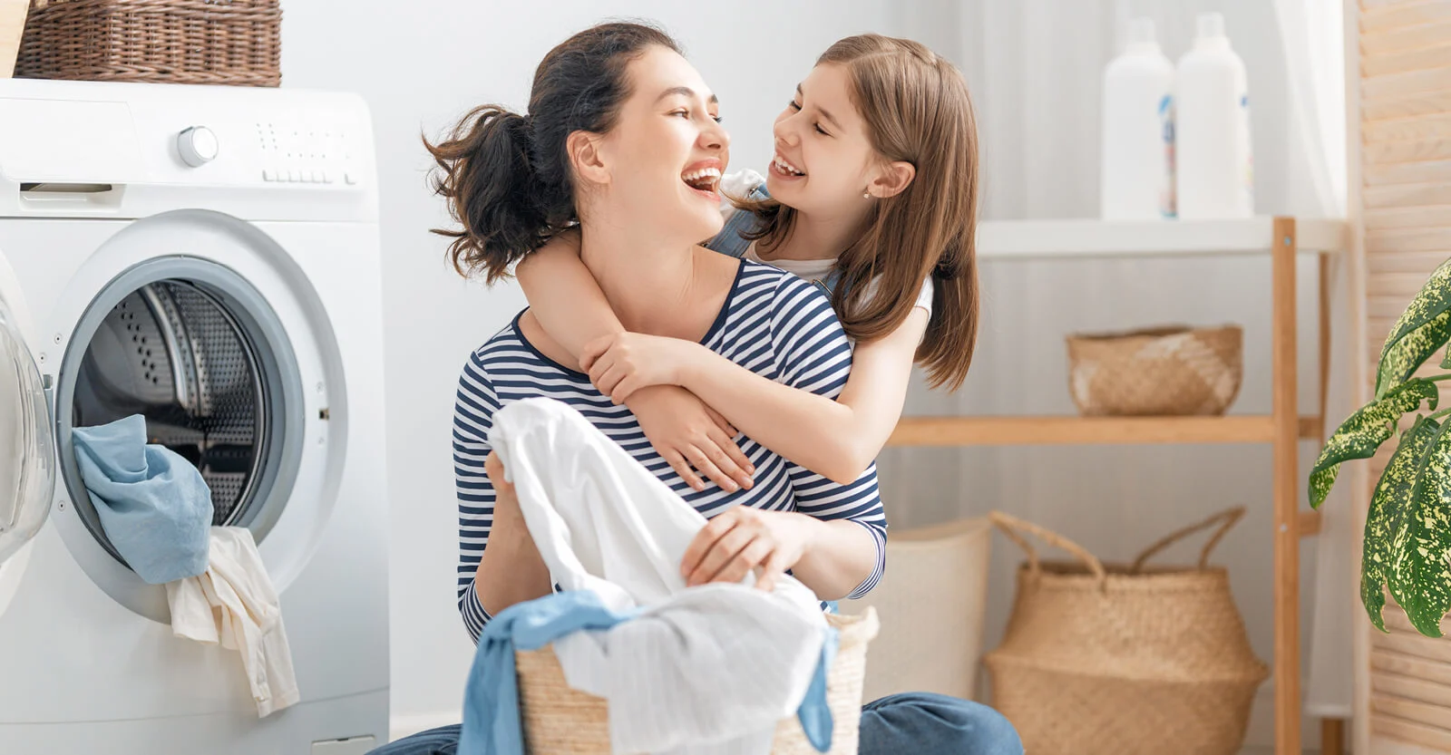 Mother and daughter laughing together while doing laundry in a bright laundry room with white washing machine and baskets.