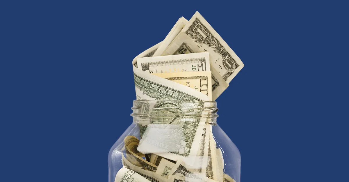 Glass jar filled with folded dollar bills against a blue background, representing savings or financial storage.