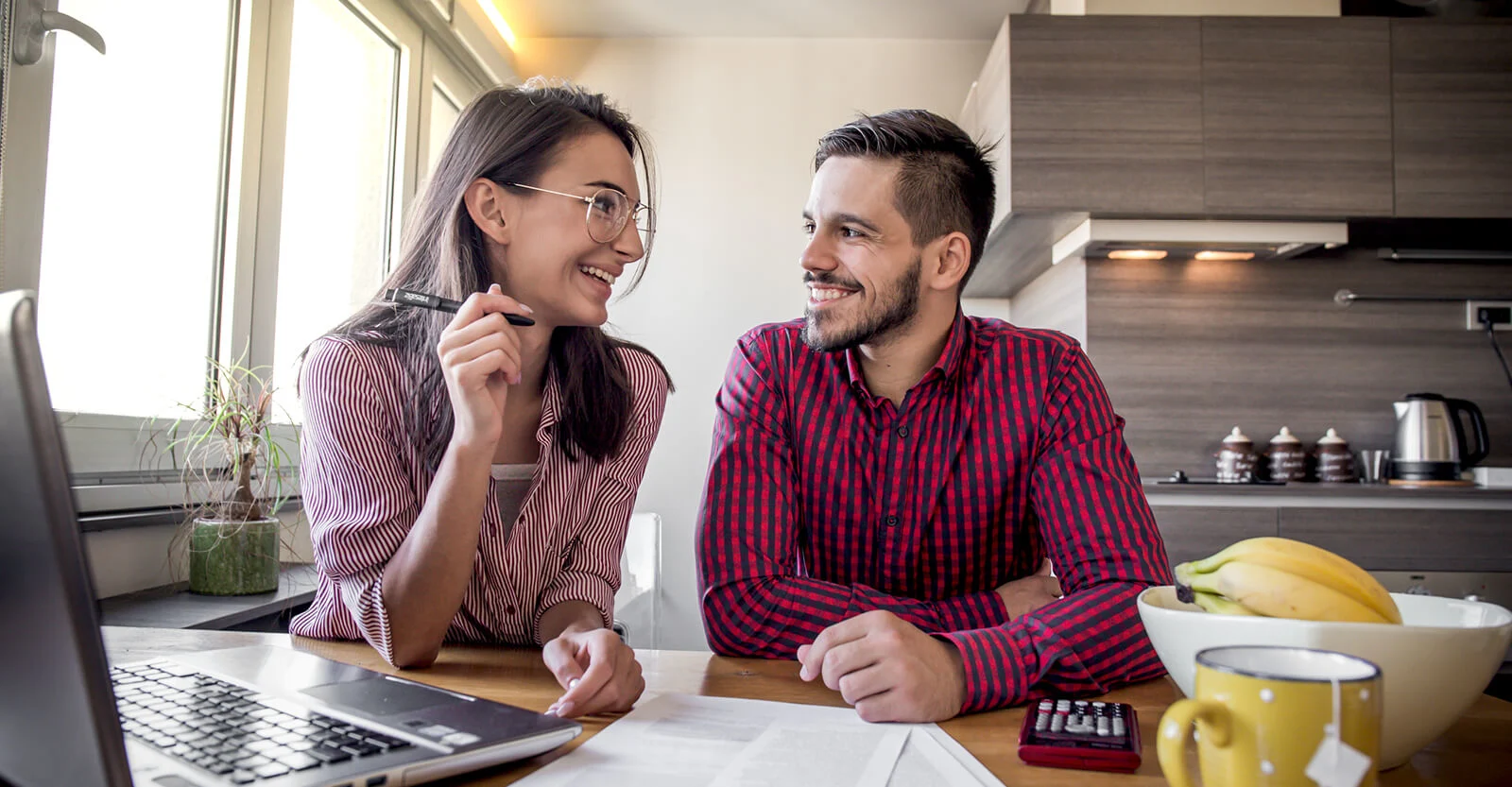 Smiling couple in striped shirts reviewing documents at kitchen table with laptop, calculator, and fruit bowl nearby.