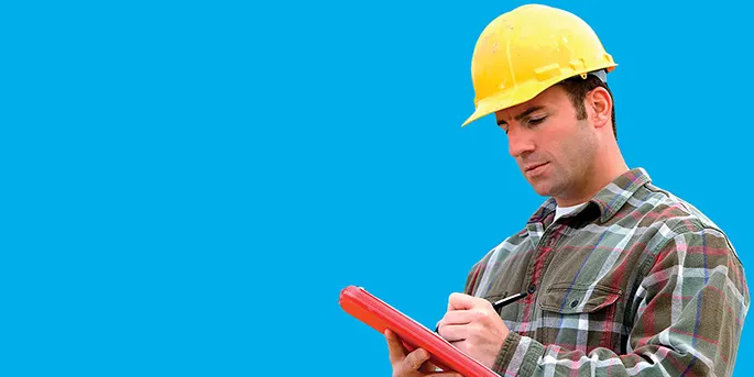 Construction worker in yellow hard hat and plaid shirt writing on red clipboard against bright blue background