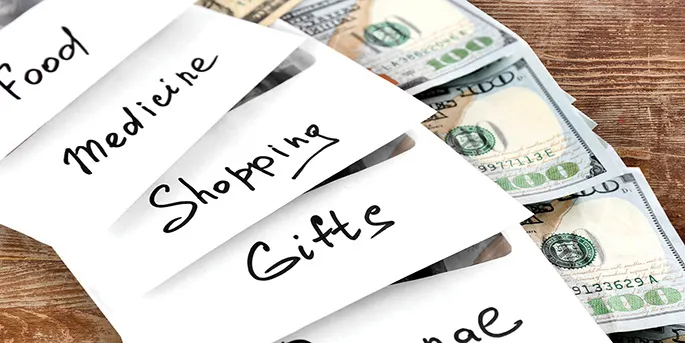 White envelopes labeled Food, Medicine, Shopping, and Gifts with hundred dollar bills inside on a wooden surface.