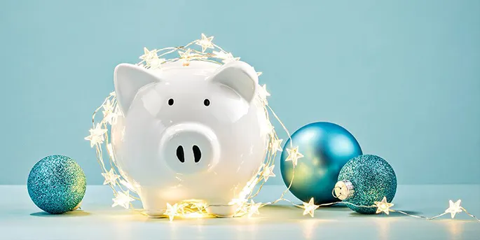 White piggy bank surrounded by glowing star-shaped string lights and blue glitter ornaments on light blue background.