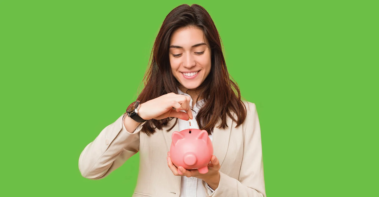 Smiling woman in beige blazer inserting coin into pink piggy bank against bright green background