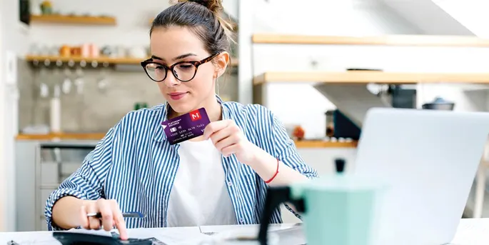 Woman in striped blazer and glasses holding credit card while working on laptop in modern kitchen setting.