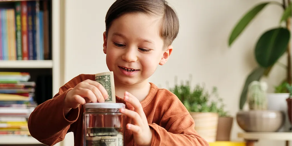Young boy in orange shirt smiling while putting a dollar bill into a clear glass jar, learning about saving money.