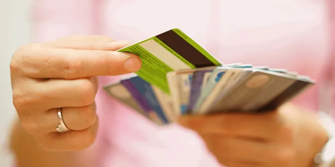 Hand holding a green credit card in front of a fanned display of multiple colorful credit cards against a soft pink background.