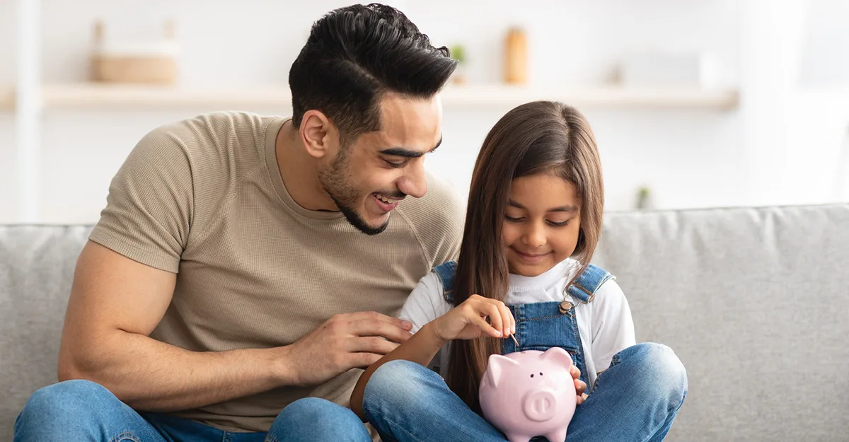 Father and daughter sitting on gray couch, smiling as the girl puts a coin into a pink piggy bank.
