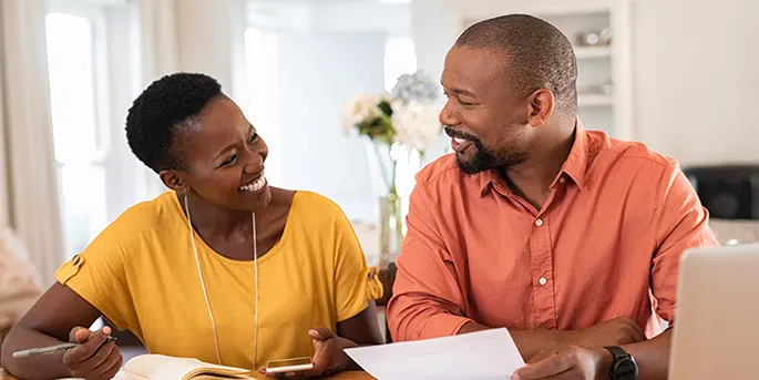 Smiling couple in yellow and orange shirts reviewing documents together at home in a bright, modern interior.