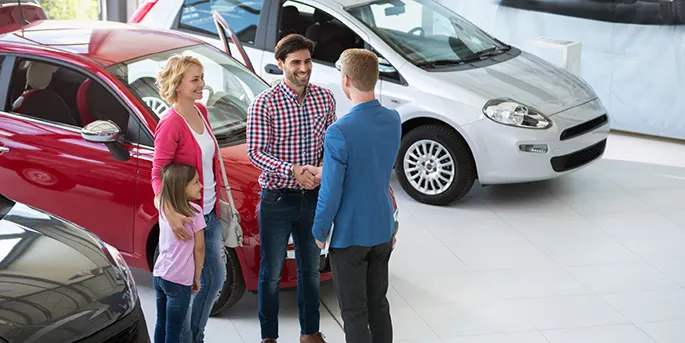 Family with young daughter meeting with car salesman in showroom, standing between red and white vehicles.