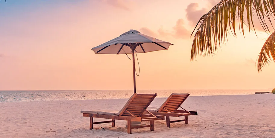 Two wooden beach loungers under white umbrella on sandy beach at sunset with palm fronds and pink-orange sky over ocean
