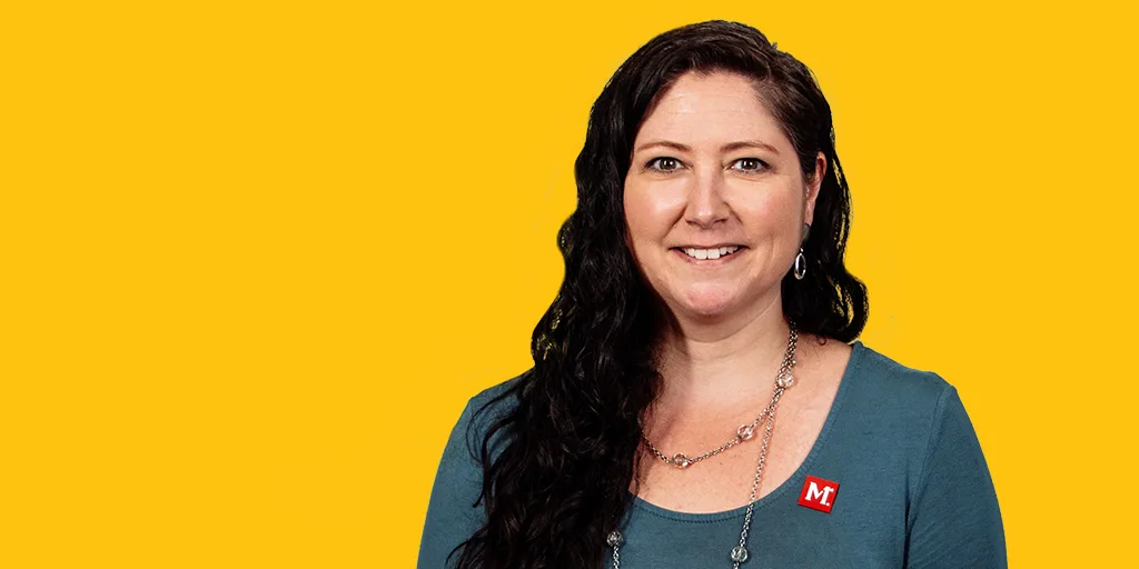 Woman with long dark hair wearing teal shirt and necklace smiling against bright yellow background.