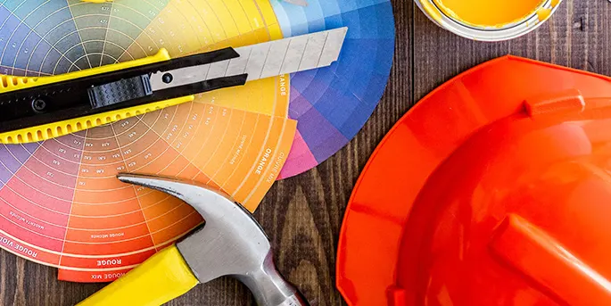 Construction tools including orange hard hat, hammer, utility knife, measuring tape, and colorful paint swatches on wooden surface.