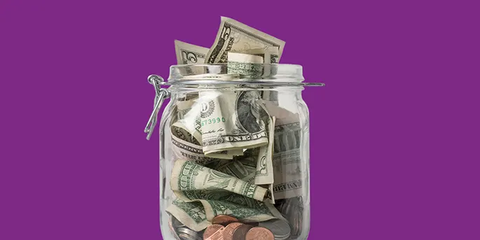 Glass jar filled with dollar bills and coins against a purple background, representing savings or money storage.