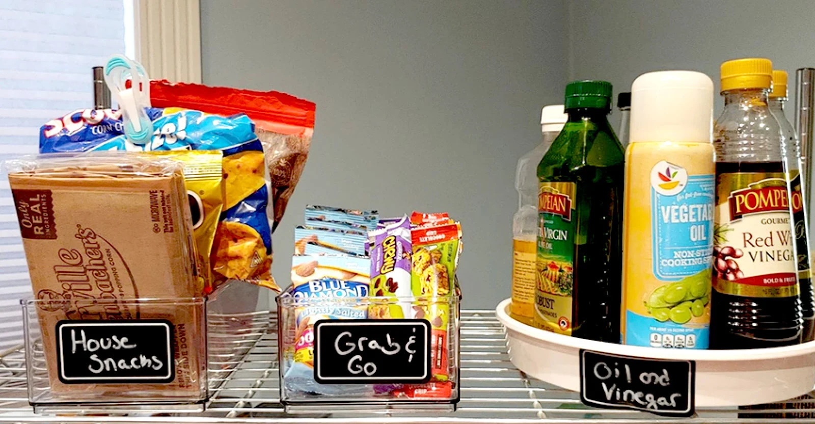 Organized pantry shelf with labeled clear containers for snacks and grab-and-go items, plus a lazy Susan with cooking oils.