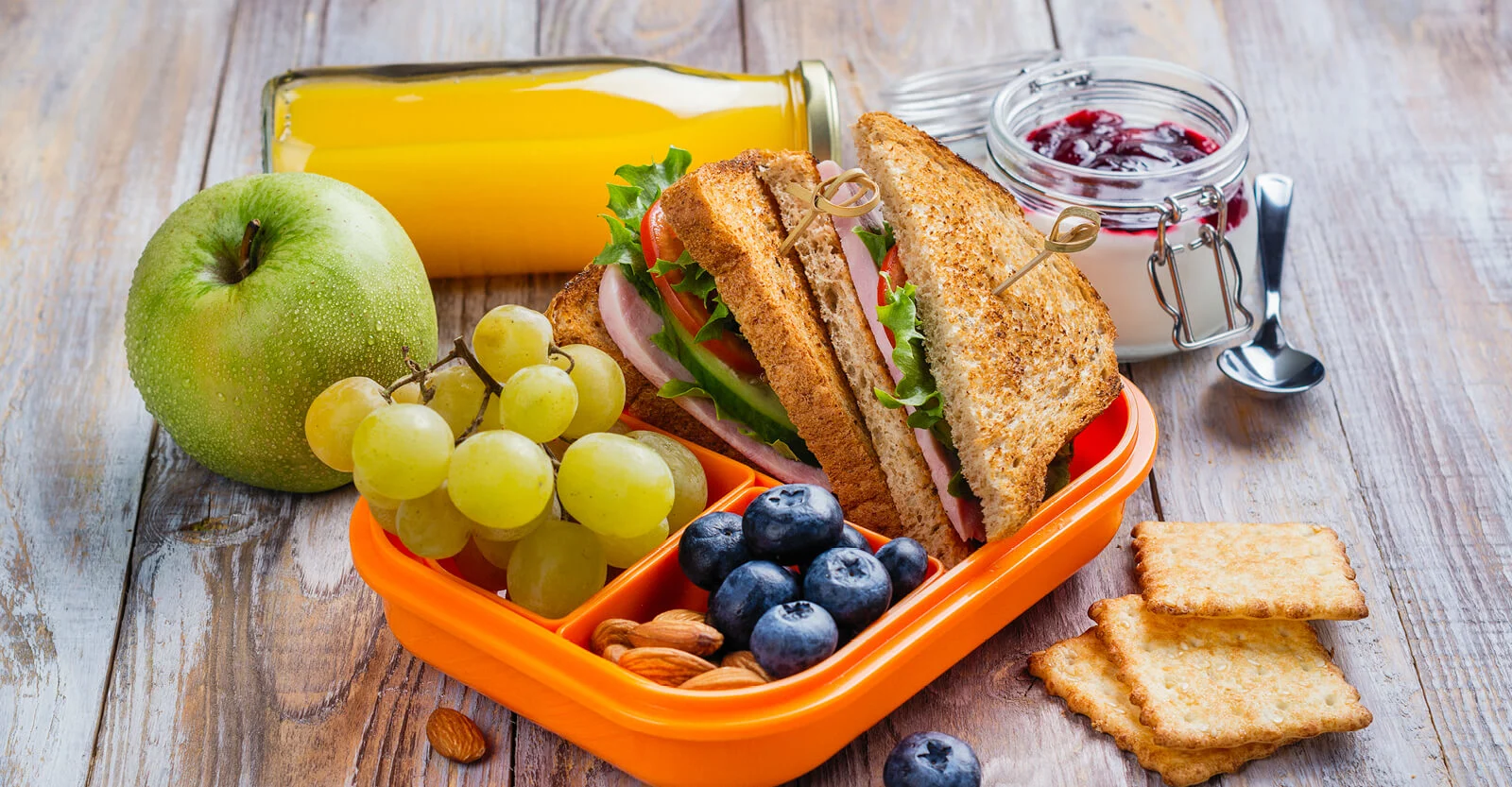 Packed lunch box with sandwich, green apple, grapes, blueberries, almonds, crackers, orange juice, and jam on wooden table.