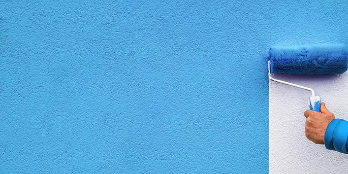 Hand holding paint roller applying blue paint to white textured wall, showing painting in progress.