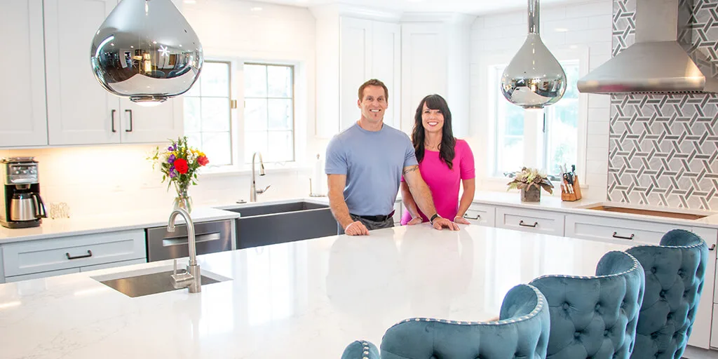 Couple standing in modern white kitchen with chrome pendant lights, white island, and blue tufted bar stools.