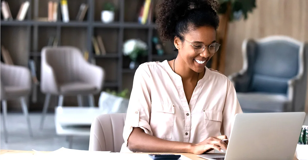 Smiling woman with glasses wearing a pink shirt working on a laptop in a modern office with bookshelves in the background.