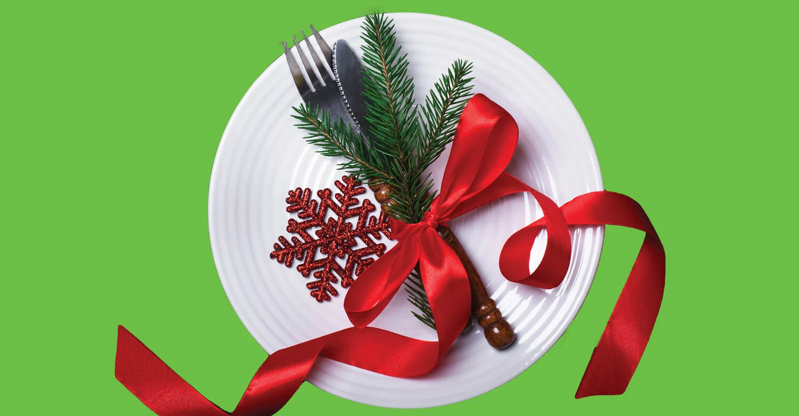 White plate with fork, knife, pine branches, red snowflake ornament, and red ribbon on green background.