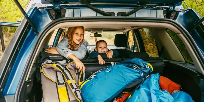 Two children smiling from the back of an open SUV filled with camping gear including blue backpacks and yellow equipment.