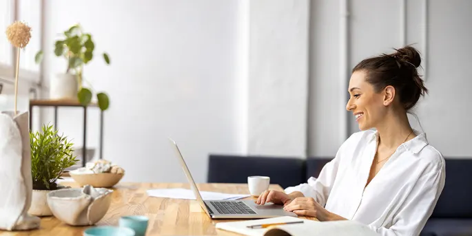 Woman in white shirt smiling while working on laptop at wooden table with coffee cup and plants in bright modern room