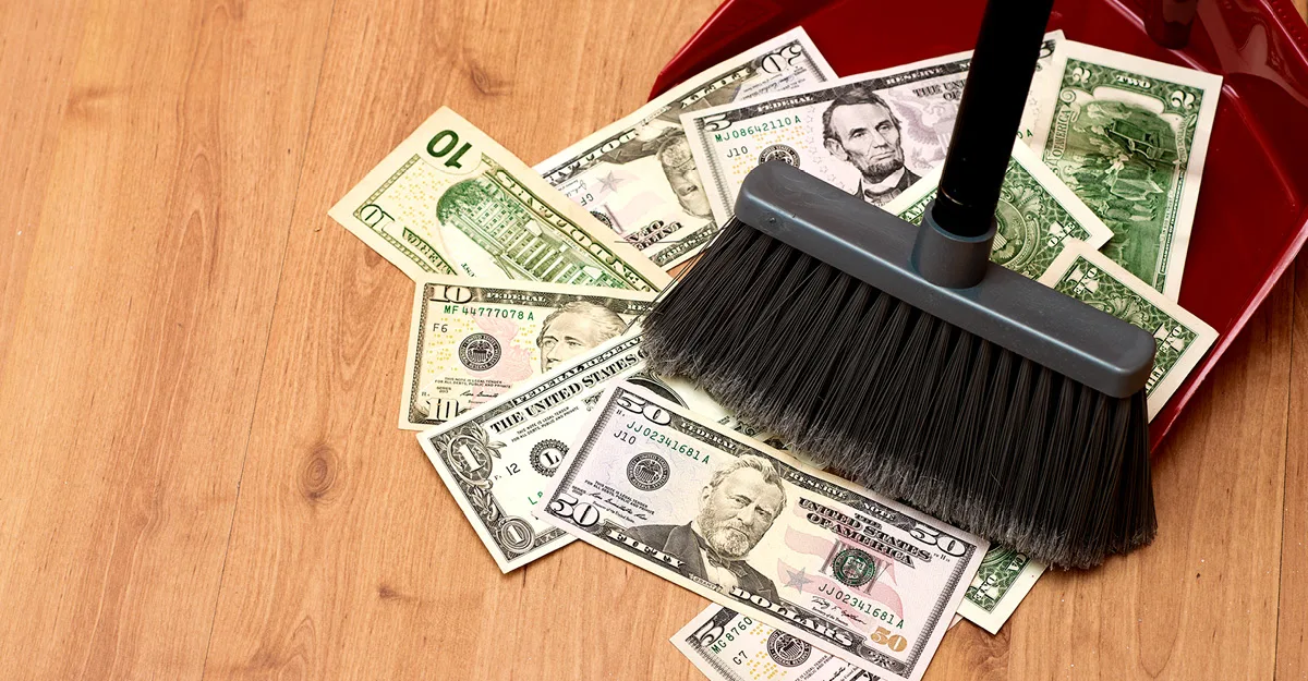 A black broom sweeping various U.S. dollar bills into a red dustpan on a wooden floor.
