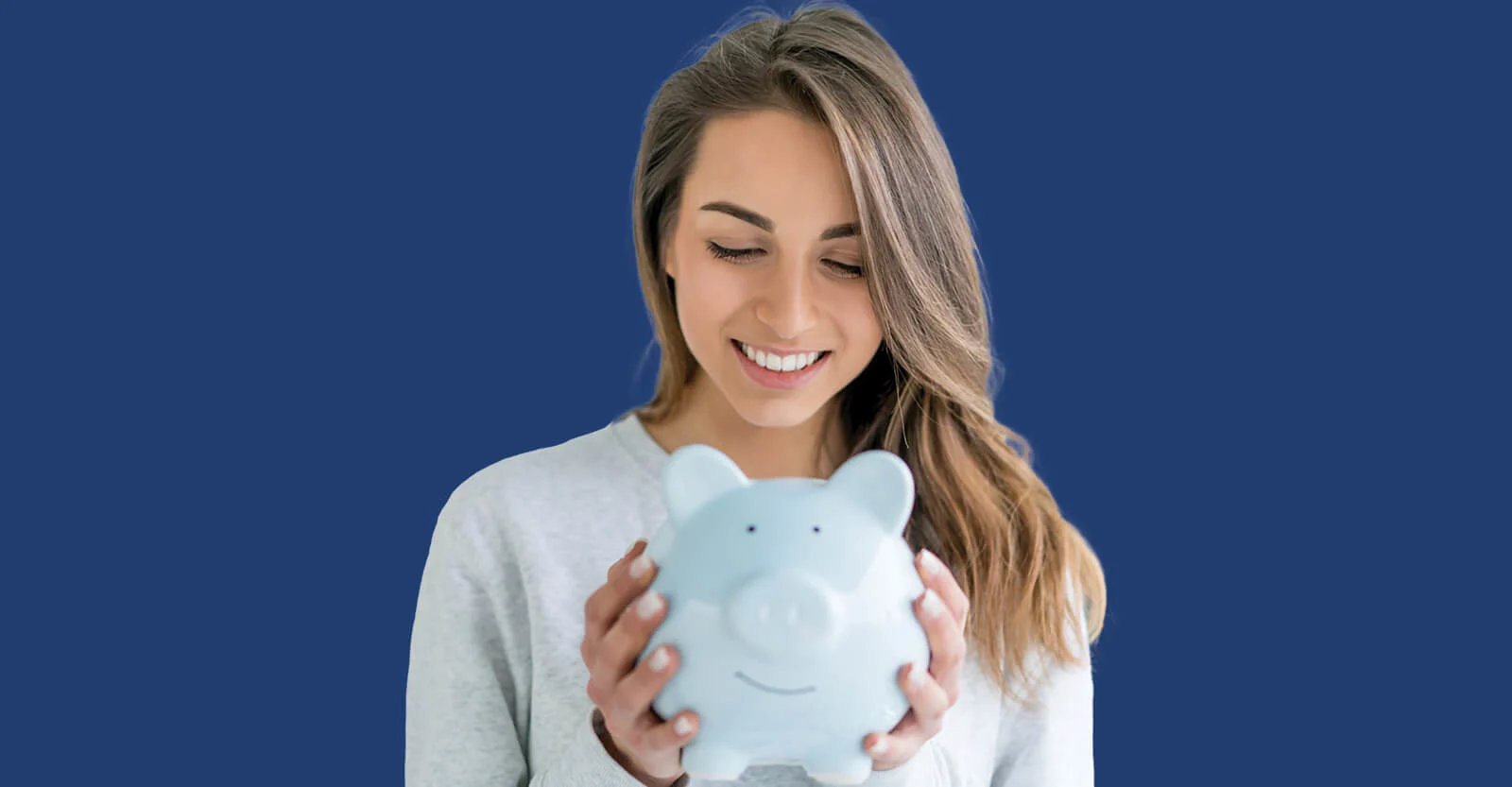 Smiling woman in white sweater holding a light blue piggy bank against a dark blue background.