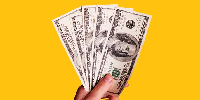 Hand holding fanned out one hundred dollar bills against a bright yellow background